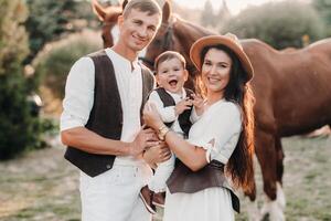 A family in white clothes with their son stand near two beautiful horses in nature. A stylish couple with a child are photographed with horses photo