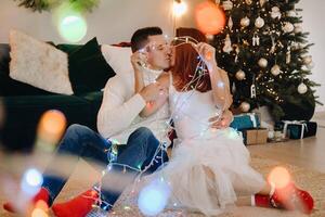 a happy married couple is sitting at home on the floor near the Christmas tree and kissing photo