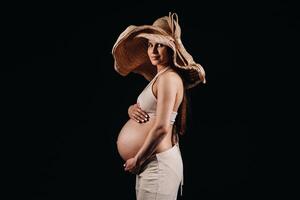 pregnant woman in a straw hat in beige clothes in the studio on a black background photo