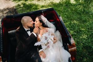 The bride and groom with a bouquet are sitting in a carriage in nature in retro style photo