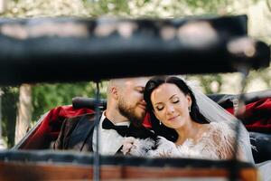 The bride and groom with a bouquet are sitting in a carriage in nature in retro style photo