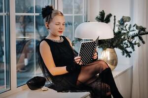 A stylish girl in a black dress sits on the windowsill by the window with a book in her hands photo
