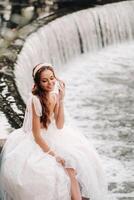 An elegant bride in a white dress, gloves and bare feet is sitting near a waterfall in the Park enjoying nature.A model in a wedding dress and gloves at a nature Park.Belarus photo