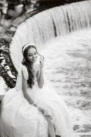 An elegant bride in a white dress, gloves and bare feet is sitting near a waterfall in the Park enjoying nature.A model in a wedding dress and gloves at a nature Park.Belarus photo