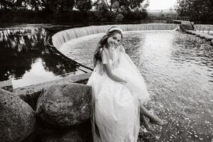 An elegant bride in a white dress, gloves and bare feet is sitting near a waterfall in the Park enjoying nature.A model in a wedding dress and gloves at a nature Park.Belarus photo