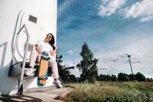 A girl in white clothes with a skate in her hands is photographed near large wind turbines in a field with trees.Modern woman with a Board for riding in a field with windmills photo