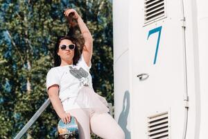 A girl in white clothes and glasses with a skate in her hands is photographed near large wind turbines in a field with trees.Modern woman with a riding Board in a field with windmills photo