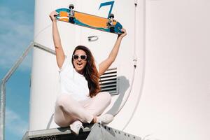 A girl in white clothes and glasses with a skate in her hands is photographed near large wind turbines in a field with trees.Modern woman with a riding Board in a field with windmills photo
