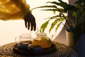 Close-up of a hand with a kettle. A teapot with sea buckthorn tea, honey and a plate with sea buckthorn on a tray photo