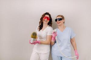 Two cosmetologists in protective glasses are holding a laser depilation device and a cactus in their hands photo