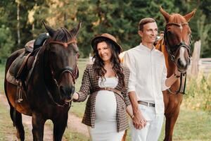 A pregnant woman in a hat with a man in white clothes walking with horses in nature. A family waiting for a child walks in the woods photo