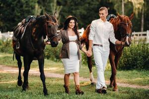 A pregnant woman in a hat with a man in white clothes walking with horses in nature. A family waiting for a child walks in the woods. photo