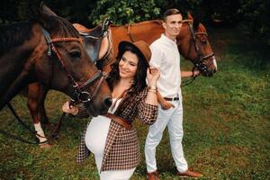 A pregnant woman in a hat with a man in white clothes walking with horses in nature. A family waiting for a child walks in the woods photo
