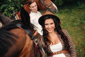 a pregnant girl in a hat and a man in white clothes stand next to horses in the forest in nature.Stylish pregnant woman with her husband with horses.Married couple. photo