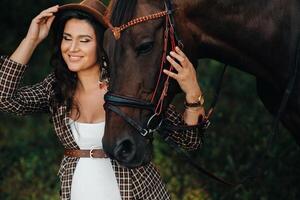 pregnant girl with a big belly in a hat next to horses in the forest in nature.Stylish girl in white clothes and a brown jacket. photo