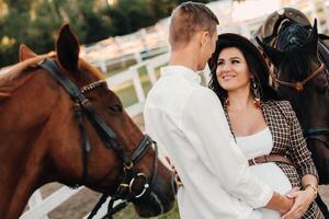 a pregnant girl in a hat and a man in white clothes stand next to horses at a White fence.Stylish family waiting for a child strolling in nature photo
