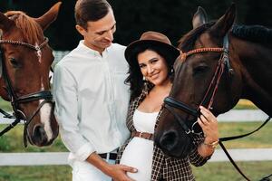 a pregnant girl in a hat and a man in white clothes stand next to horses at a White fence.Stylish family waiting for a child strolling in nature photo