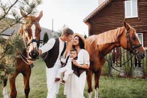A family in white clothes with their son stand near two beautiful horses in nature. A stylish couple with a child are photographed with horses photo