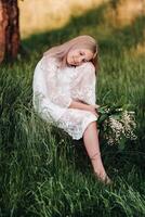 A beautiful nine-year-old blonde with long hair in a long white dress, holding a bouquet of lilies of the valley, sits on a stone in the Park.Summer, sunset. photo