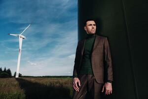 A man in a business suit with a green Golf shirt stands next to a windmill against the background of the field and the blue sky.Businessman near the windmills.Modern concept of the future. photo