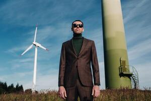 A man in a business suit with a green Golf shirt stands next to a windmill against the background of the field and the blue sky.Businessman near the windmills.Modern concept of the future. photo