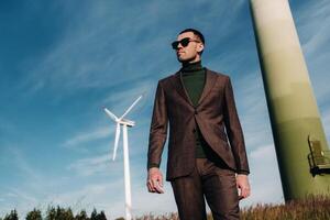 A man in a business suit with a green Golf shirt stands next to a windmill against the background of the field and the blue sky.Businessman near the windmills.Modern concept of the future. photo