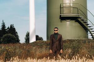A man in a business suit with a green Golf shirt stands next to a windmill against the background of the field and the blue sky.Businessman near the windmills.Modern concept of the future. photo