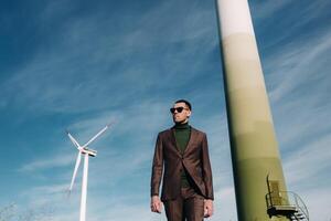 A man in a business suit with a green Golf shirt stands next to a windmill against the background of the field and the blue sky.Businessman near the windmills.Modern concept of the future. photo