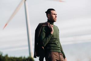 A man in a business suit with a green Golf shirt stands next to a windmill against the background of the field and the blue sky.Businessman near the windmills.Modern concept of the future. photo