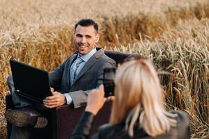 Portrait of a businessman in a suit with a laptop sitting in a chair in a wheat field and photographed by an unrecognizable girl. photo