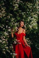 a girl in a red dress with red lips stands next to a large white flowering tree At sunset photo