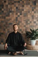 Portrait of a young man in a black kimano sitting in a lotus position on a gym mat in the interior photo