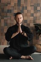 Portrait of a young man in a black kimano sitting in a lotus position on a gym mat in the interior photo