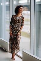 a brunette woman in a tiger dress stands near a large window photo