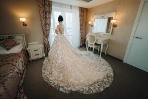 The bride in a beautiful dress with a long train stands near the window in the interior. photo