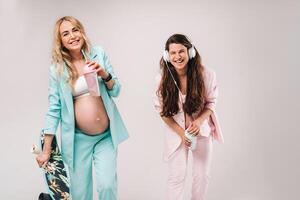 Two pregnant girls in turquoise and pink suits with glasses of juice, a skate and headphones stand on a gray background photo