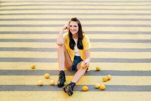 a girl with lemons in a yellow shirt, shorts and black shoes sits on a yellow pedestrian crossing in the city. The lemon mood photo