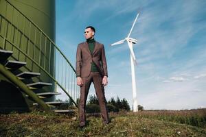 A man in a business suit with a green Golf shirt stands next to a windmill against the background of the field and the blue sky.Businessman near the windmills.Modern concept of the future. photo