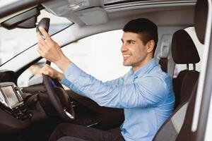 Young Driver. Happy Guy Checking Mirrors Sitting In Driver's Seat Testing Buying Auto In Dealership Store, Side View photo