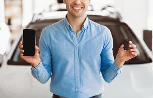 Happy man showing new car key and smartphone with blank screen standing in automobile dealership center, mock up photo