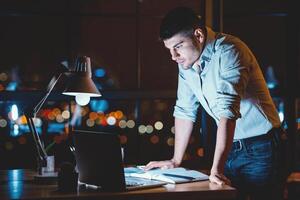 Serious Entrepreneur Man Standing Looking At Laptop Working Standing In Modern Office With Panoramic View At Night. Copy Space photo