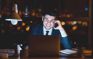 Happy Business Guy Having Phone Conversation On Cellphone Sitting At Laptop Overnight In Office. Corporate Lifestyle And Communication photo