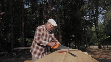 A senior craftsman focuses on cutting wood with precision while surrounded by lush trees under a bright afternoon sun. The tranquility of nature enhances the moment. video