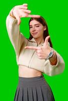 A young woman stands against a vibrant green screen, creating a finger frame with her hands as if visualizing a composition or focusing on a specific view. photo