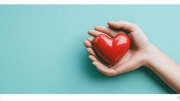A hand gently cradles a shiny red heart on a serene teal background, symbolizing love and compassion in a simple yet profound moment photo