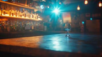 A glass sits on the bar counter in a dimly lit bar with illuminated shelves of liquor behind it during the evening photo