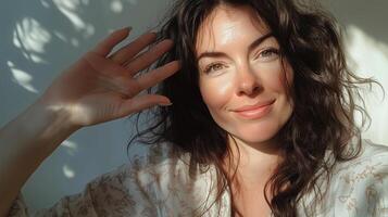 A woman with wavy hair smiling and waving near a sunny window with playful shadows in the background during the afternoon photo