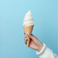 A hand holding a swirl of soft serve ice cream in a cone against a light blue backdrop during a sunny day photo