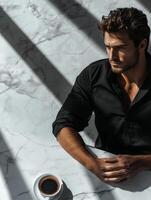 A man in a black shirt sits contemplatively at a marble table enjoying a cup of coffee in soft sunlight photo