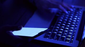 A person types on a laptop with one hand while holding a smartphone in the other, illuminated by a faint blue light. video